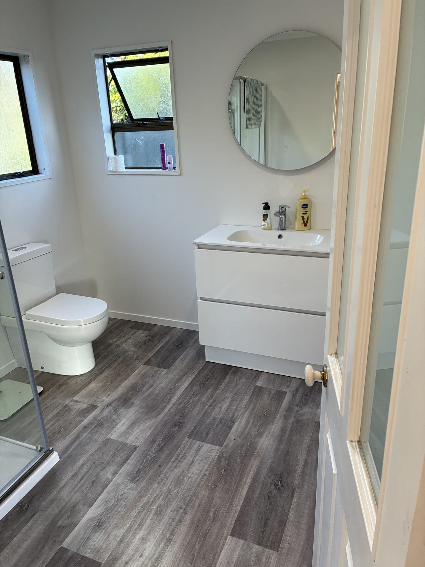 Completed bathroom renovation with a white vanity, round mirror and grey timber-look flooring
