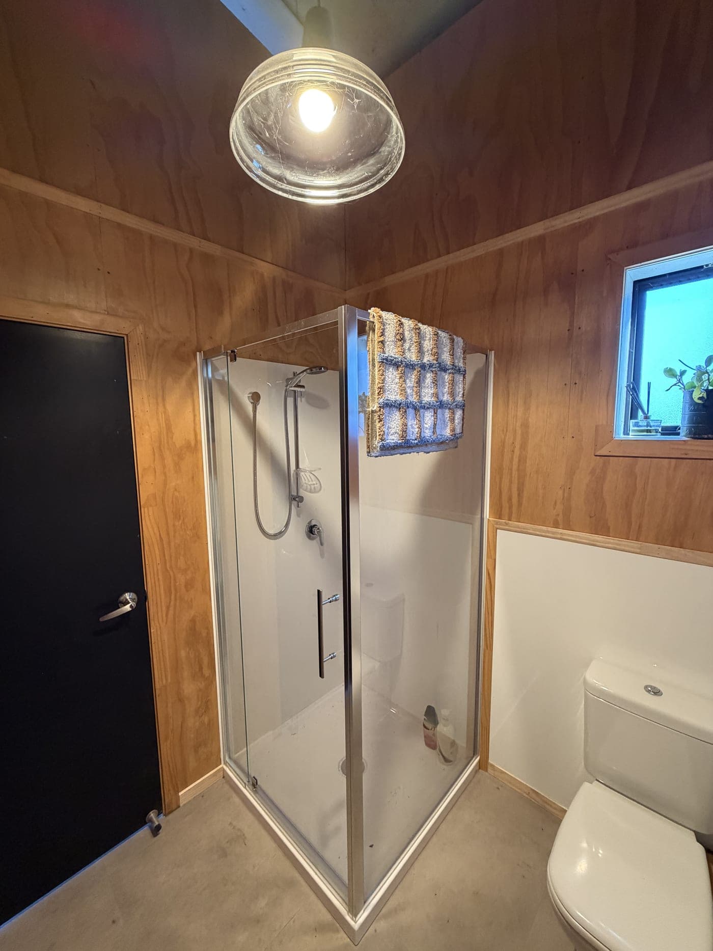 Corner glass shower in the pole shed bathroom, with ply-lined walls and a small window