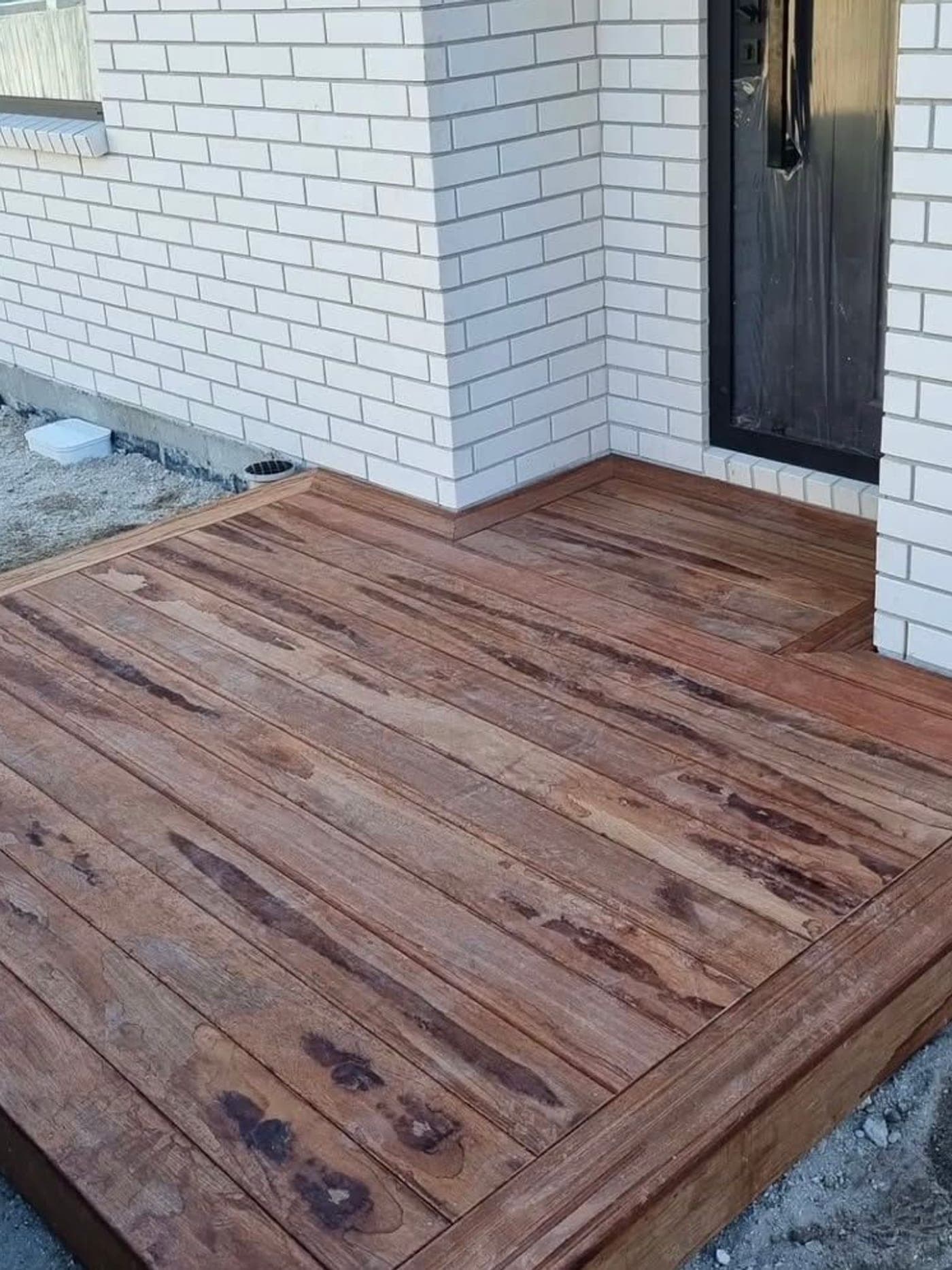 Hardwood timber entrance deck wrapping the corner of a white-brick house and meeting a black front door